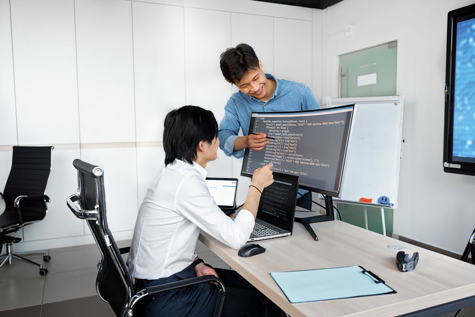 Two young professionals collaborating at a desk with laptops and monitors in a modern office setting